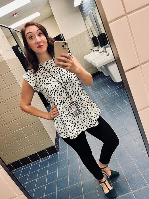 A reviewer captures a mirror selfie in a restroom, donning a black-and-white polka-dot blouse paired with black pants, accessorized with an ID badge and phone lanyard