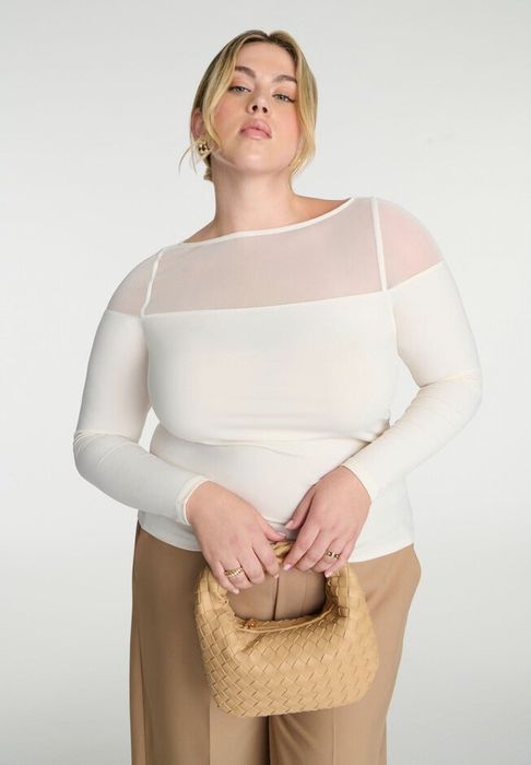 Person wearing a sheer-paneled long-sleeve top and holding a woven tan handbag, posing for a shopping feature