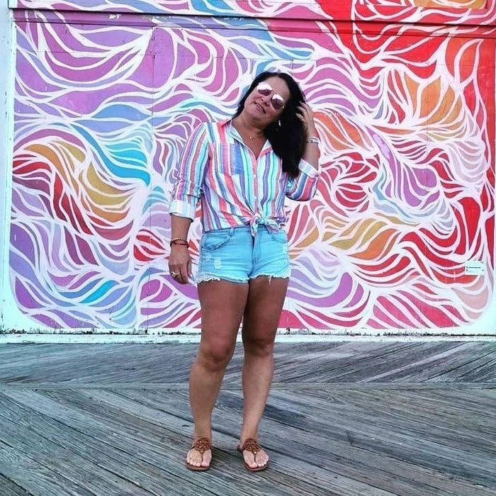 Reviewer in sunglasses, a pastel striped shirt, and denim shorts posing in front of a vibrant mural with wavy designs