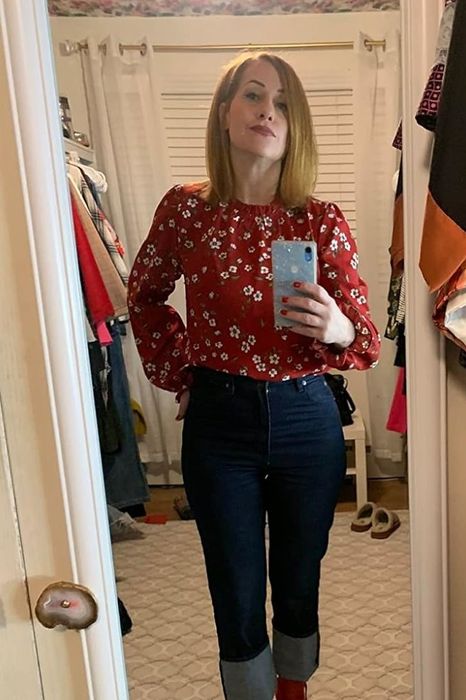 A person snapping a mirror selfie in a floral top and cuffed jeans, surrounded by vibrant clothing in a closet