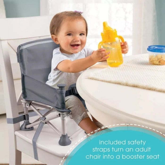 A toddler seated in a portable booster seat at a table, secured with safety straps and holding a yellow bottle