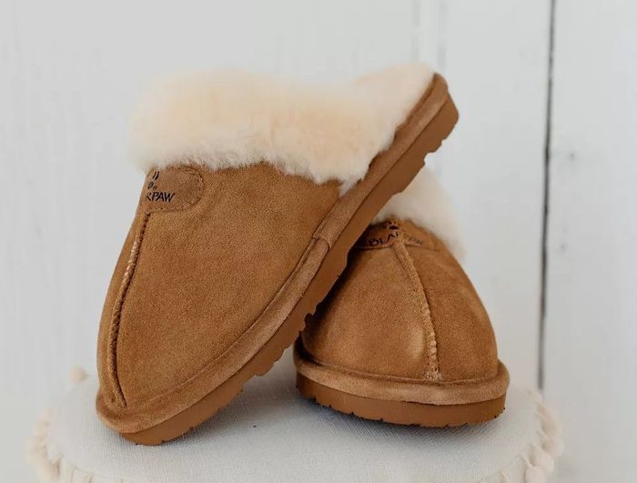 A pair of suede and faux fur slippers, one standing upright and the other resting against it, showcased on a white surface with pom-pom detailing