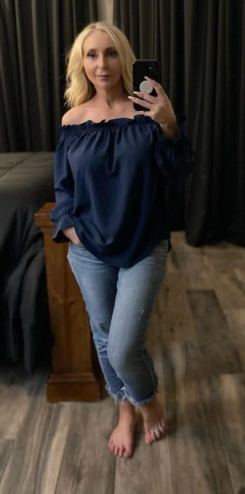 Person capturing a mirror selfie in an off-the-shoulder top and jeans, standing on a wooden floor with a dark background