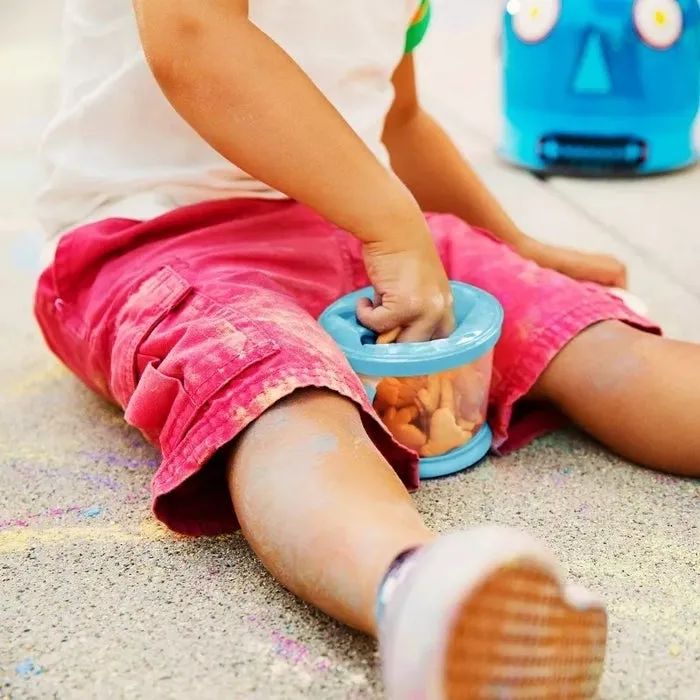 A child sitting on the floor, playing with a toy and reaching into a container, with a toy robot placed nearby