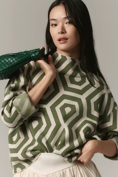 Model posing in a geometric-patterned sweater, holding a woven green bag, showcasing a fashion-forward look for a shopping feature