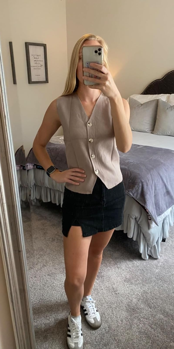 Individual snapping a mirror selfie in a sleeveless buttoned top, paired with a black skirt and sneakers, standing in a bedroom with a tidy bed