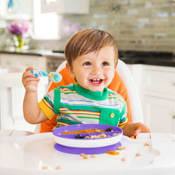 A cheerful toddler in a high chair, wearing a striped shirt, holds a spoon over a bowl filled with food and berries in a cozy kitchen environment
