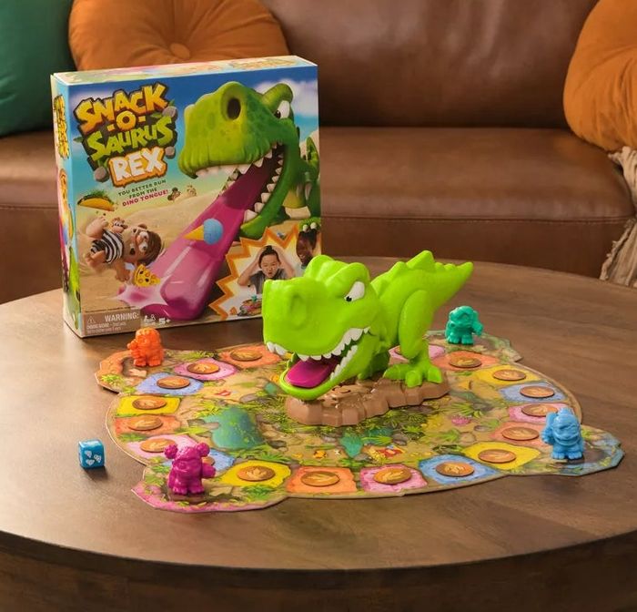 A board game titled Snack Attack! Dinosaur Rex displayed on a coffee table with game pieces, dice, and the game box. The background shows sofas and cushions arranged randomly