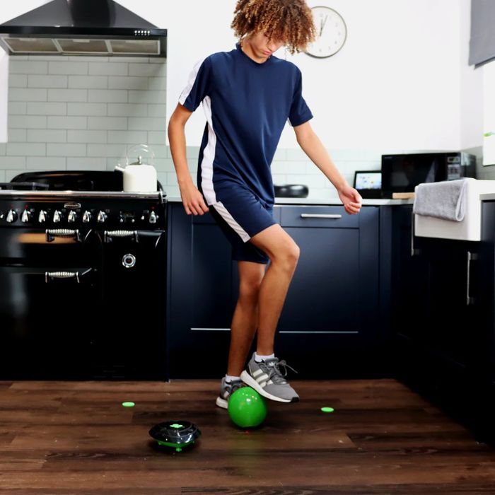 A teenager in sportswear practicing soccer in a contemporary kitchen, with a small robot vacuum nearby