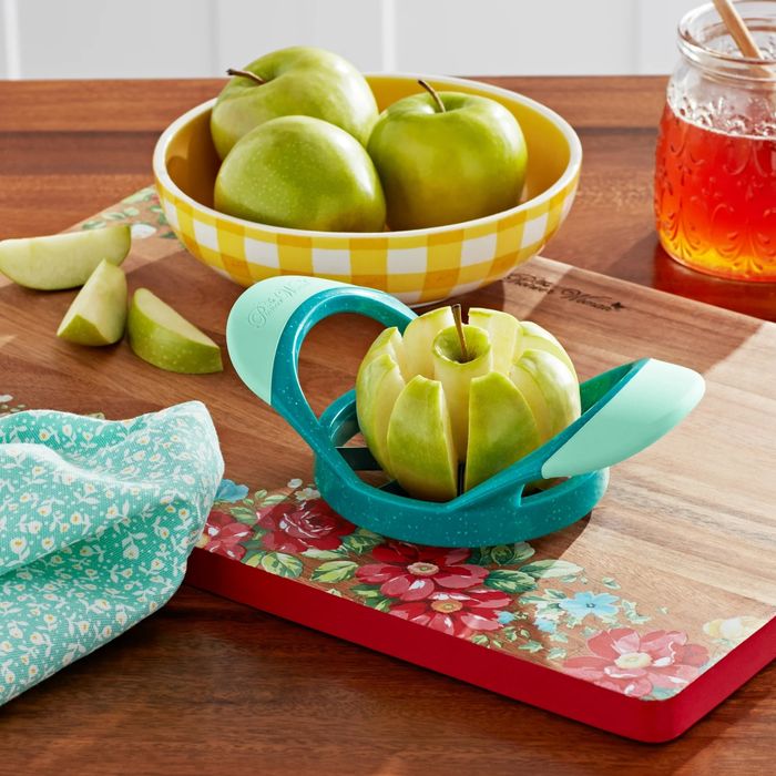 An apple slicer cutting a green apple on a decorative cutting board, with whole apples in a bowl and a jar of honey nearby