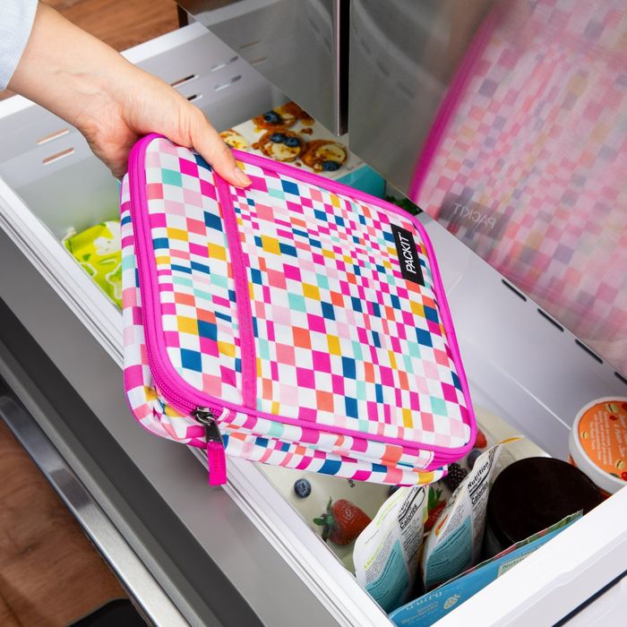 A person placing a vibrant, checkered lunch bag into a freezer drawer filled with groceries. The bag is designed by PackIt