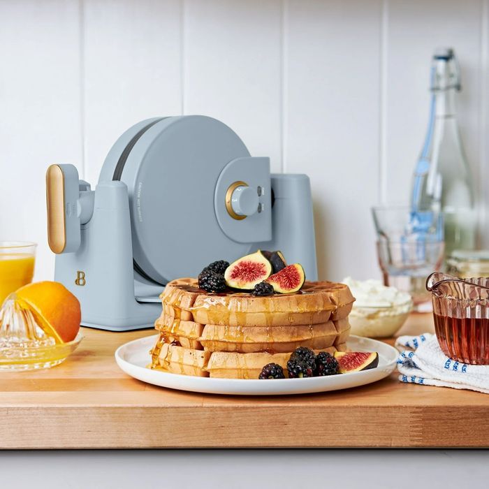 A waffle maker placed on a kitchen counter, surrounded by waffles, figs, blackberries, orange juice, cream, and syrup, setting a warm and inviting breakfast atmosphere