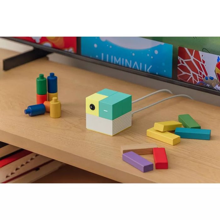 Vibrant geometric blocks and a compact cube-shaped electronic device displayed on a wooden shelf, surrounded by colorful children’s toys