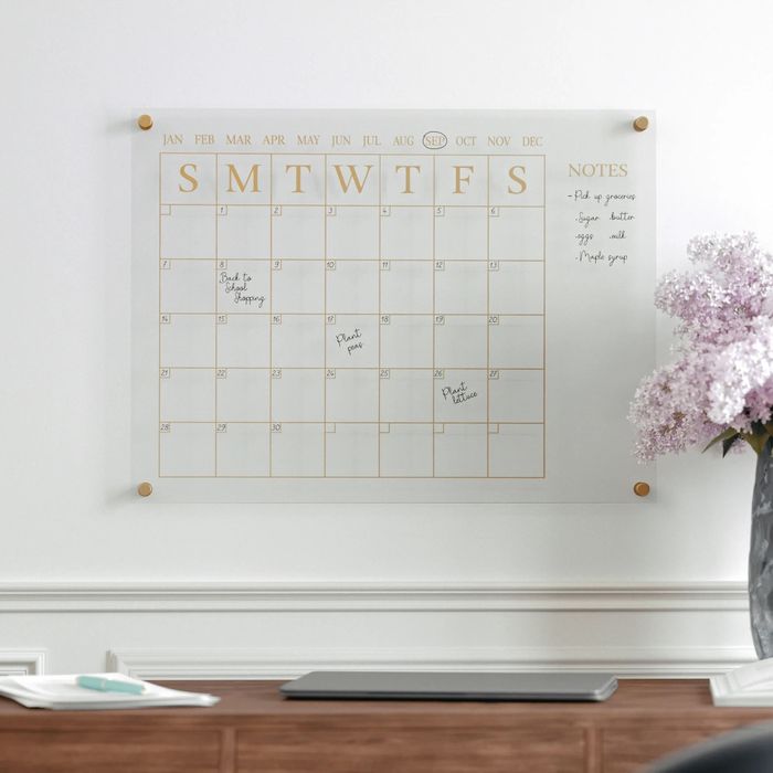 A glass wall calendar mounted above a desk, filled with handwritten notes and reminders for the month, alongside a laptop and a vase of flowers