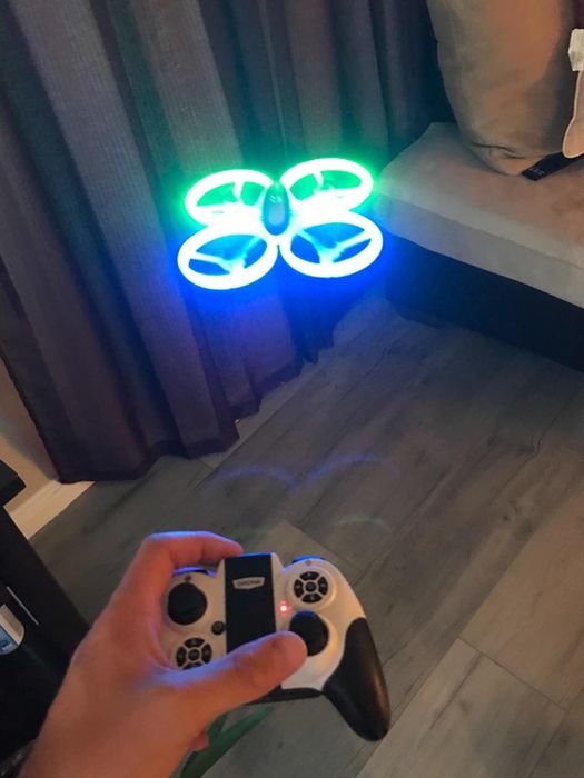 A hand holding a white and black remote, controlling a drone with blue and green LED lights.