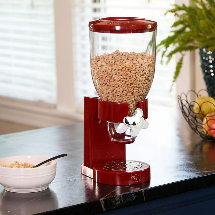 A cereal dispenser placed on a kitchen counter alongside a bowl and a fruit basket