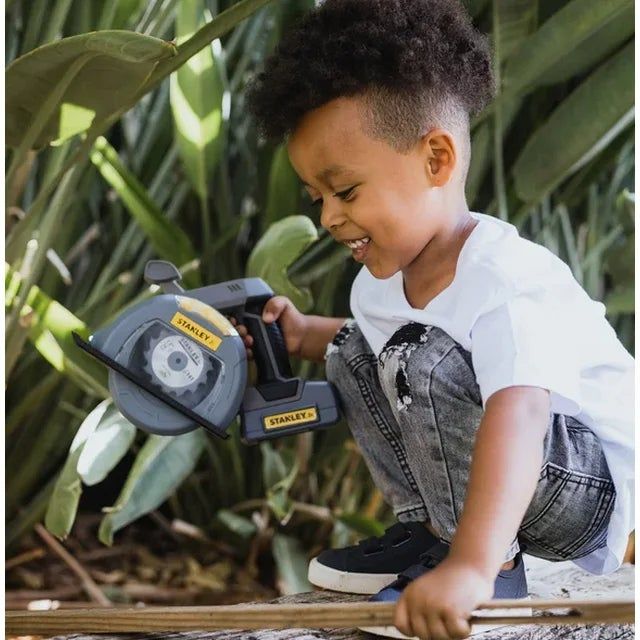 A child dressed casually smiles while crouching outdoors, holding a toy saw, capturing a joyful moment in a garden environment