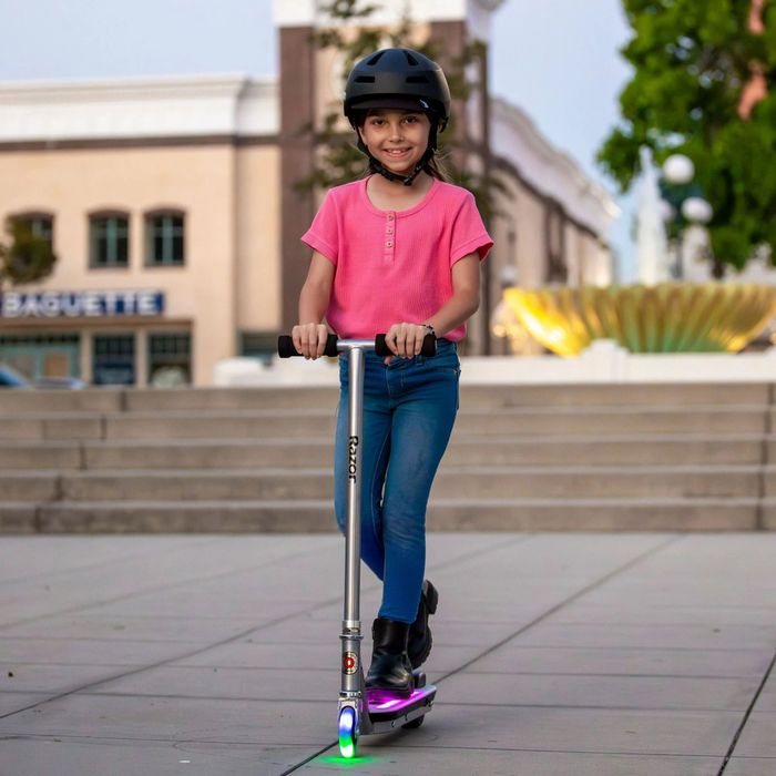 A child rides a scooter with illuminated wheels, wearing a helmet and casual attire, cruising down a city street