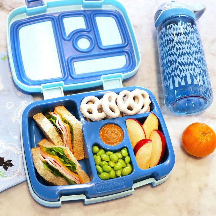 A bento lunchbox containing a sandwich, apple slices, yogurt pretzels, edamame, and a dip compartment, placed beside a water bottle and an orange on a sleek marble countertop