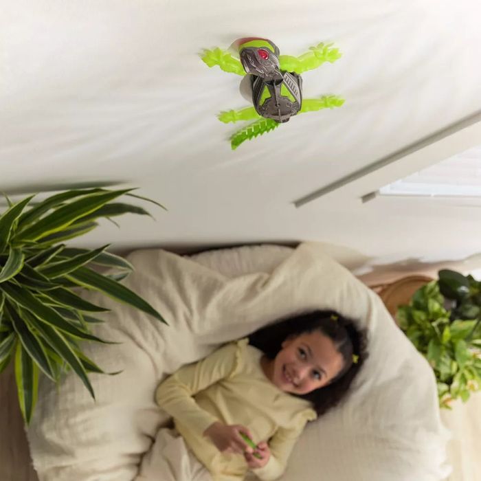 A girl lies on her bed, smiling as a robotic lizard toy crawls on the wall above her. Plants are visible in the background
