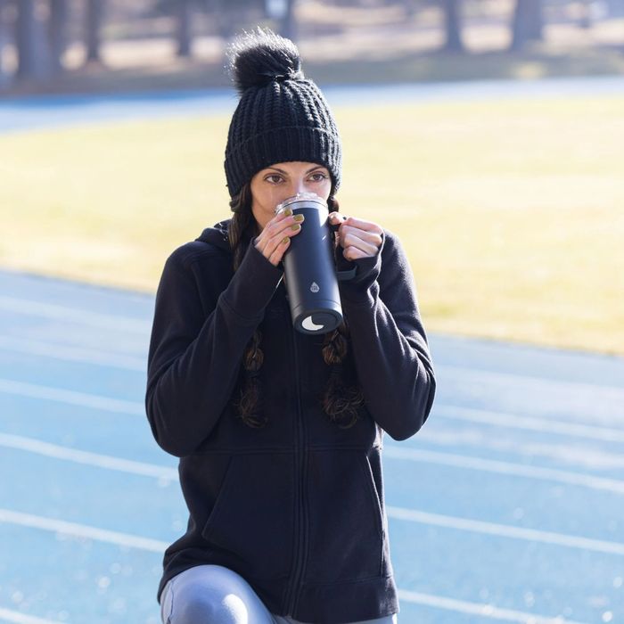A woman in a knit hat and hoodie enjoys a drink from a travel mug while on a track or field, evoking a sporty and casual outdoor vibe