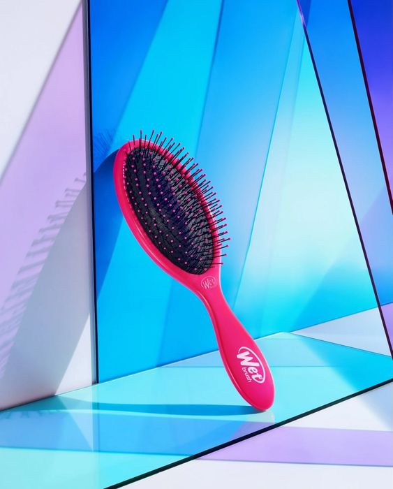 A hairbrush with black bristles on a pink base, positioned at an angle against geometric panels, showcased in a shopping article