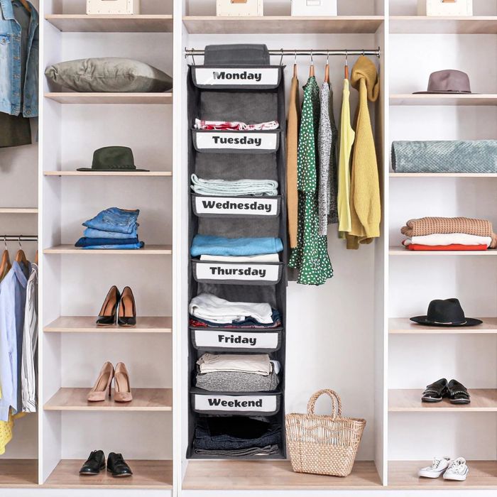 A well-organized closet featuring labeled hanging shelves for each day of the week, neatly folded clothes, shoes, hats, and a basket
