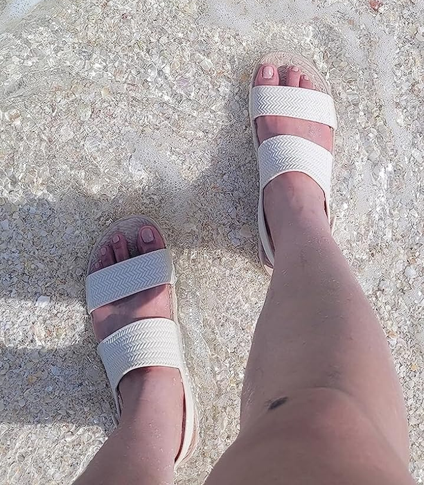 Person standing on a sandy beach, wearing white slide sandals near the shoreline