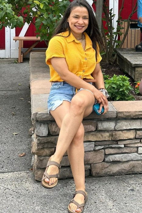The reviewer sits on a stone ledge, dressed in a yellow shirt, denim shorts, and sandals, smiling at the camera