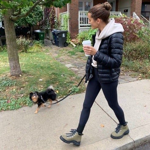 A woman in a puffy jacket and green boots walks her small dog while holding a coffee cup.