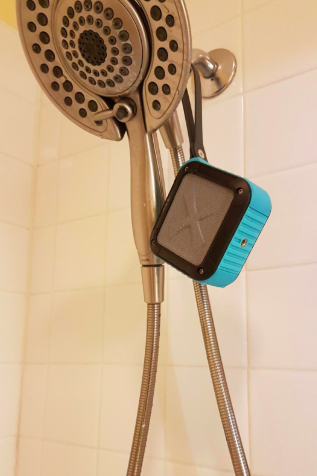 The blue, black, and gray square speaker hanging by its strap from a showerhead.