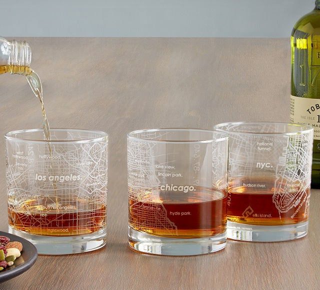 Three lowball glasses engraved with maps of LA, Chicago, and NYC, each filled with bourbon.