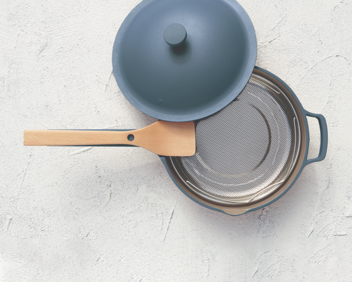 The blue pan with its lid removed, showcasing the steamer basket inside and the wooden spatula resting on the extended handle.