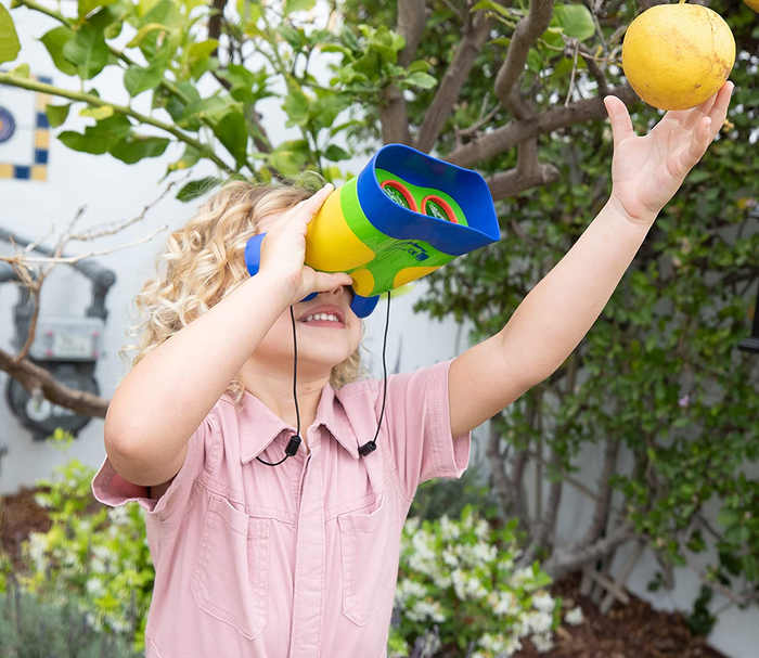 A child peering at an apple hanging from a tree through a pair of kid-sized binoculars.
