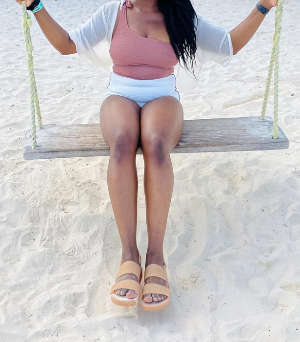 A reviewer in a casual outfit with a tank top, shorts, and sandals sitting on a wooden swing on a sandy beach