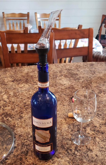A blue wine glass rests on a granite countertop, with a glass aerator on top adding about three and a half inches to its height. It sits beside an empty wine glass, with a dining table in the background.