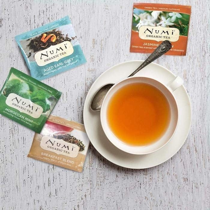 A photo featuring a white saucer with a teacup filled with vibrant orange tea, accompanied by a spoon, and four colorful tea packets placed beside the saucer.