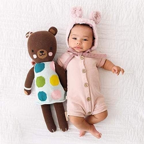 A baby sitting next to an adorable brown teddy bear featuring pink cheeks and dressed in a polka dot outfit