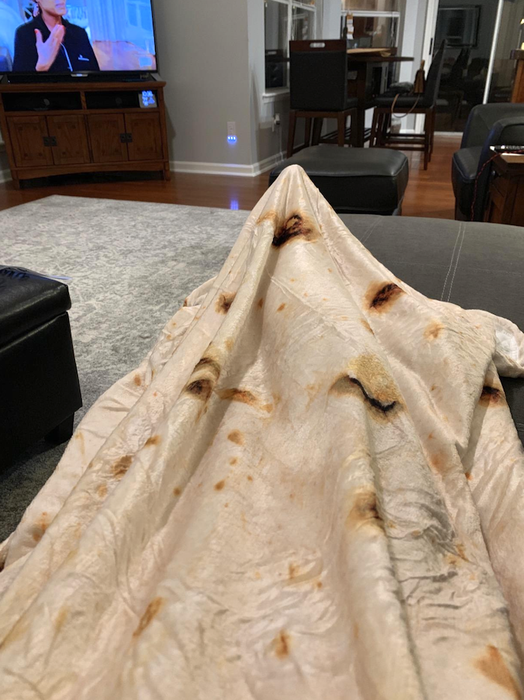 The blanket is designed to resemble an oversized burrito, with cream-colored fabric and dark spots that mimic those created when burritos are cooked. It's draped over a person’s legs, who is lounging on a chair and watching television.