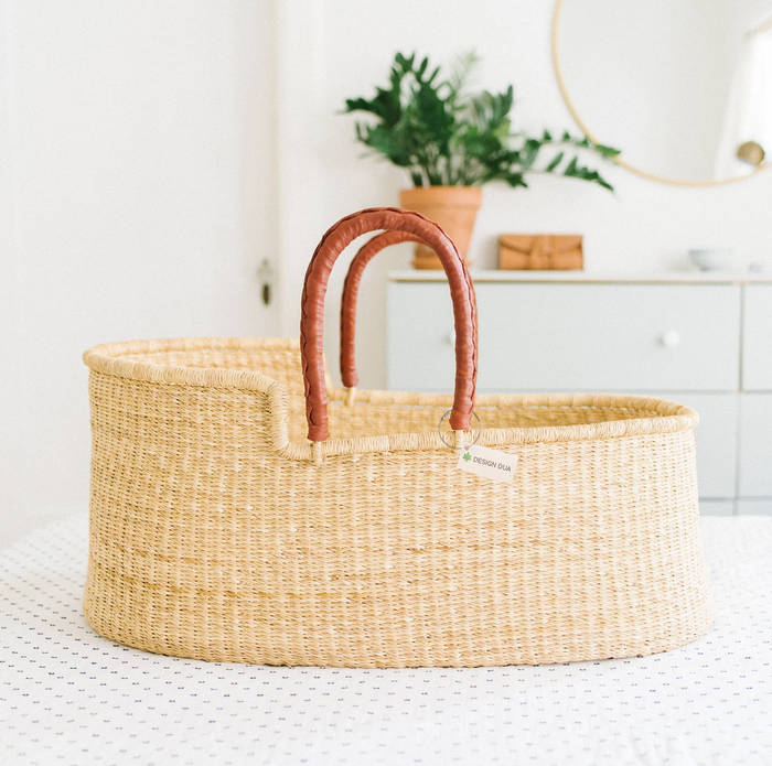 A light-colored woven Moses basket featuring leather-covered handles on both sides