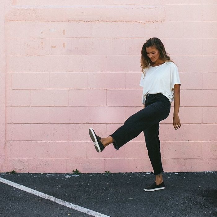 A model wearing black sneakers in front of a pink wall, playfully kicking their feet.