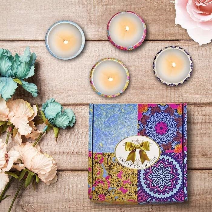 a vibrant gift box placed next to four candles in tin cans, each lit and featuring designs that complement the box