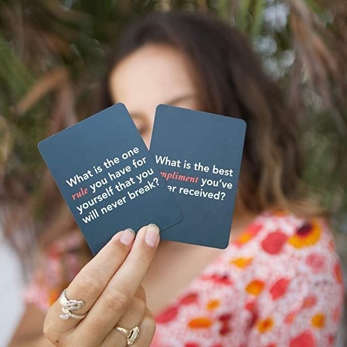 A model holding two game cards with questions: 'What is one rule you have for yourself that you will never break?' and 'What is the best compliment you’ve ever received?'