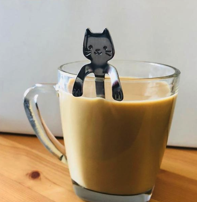A glass coffee mug sitting on a wooden countertop, filled with coffee. The cat-shaped spoon rests on top, with the cat's head peeking over the rim while its paws hang down to support the spoon.