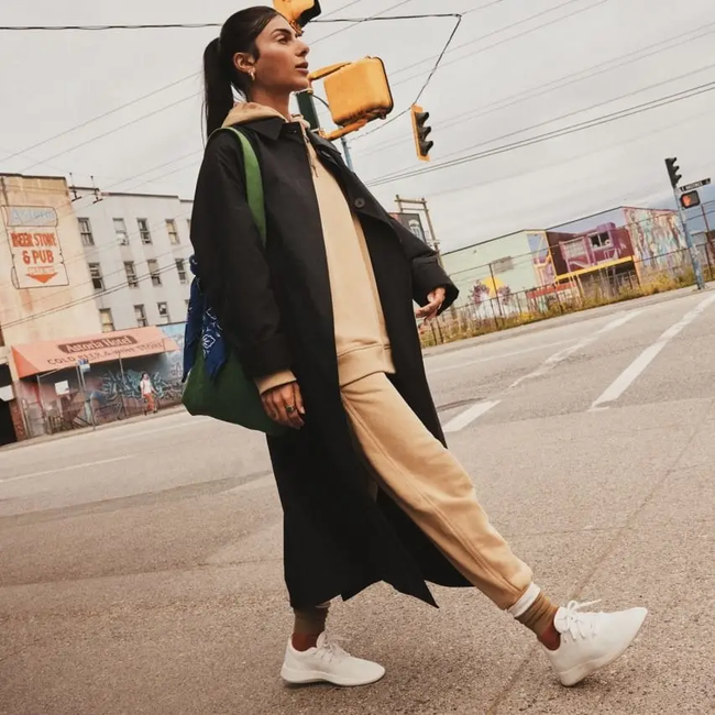 A woman dressed in layers with a coat, holding a green bag, strolling along the street