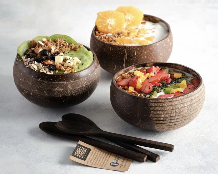 Three coconut bowls filled with smoothie bowls, accompanied by spoons placed on a table