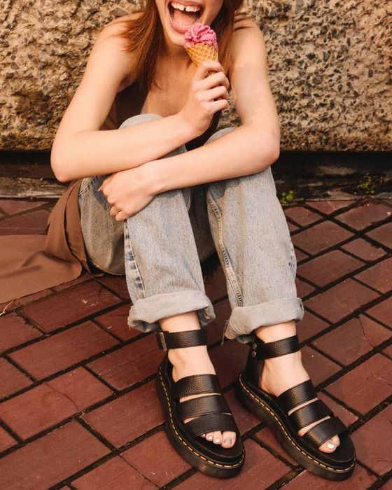 A model sitting on the pavement, laughing while holding an ice cream cone, wearing Dr. Martens sandals and denim.