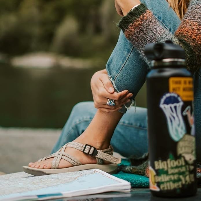 A person sitting with a map and water bottle, dressed in a striped sweater, denim jeans, and strapped sandals.
