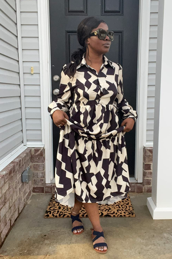 A reviewer wearing a patterned dress and sunglasses stands by a door.
