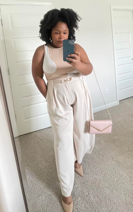 reviewer in a sleeveless top and high-waisted pants with a belt, capturing a mirror selfie while holding a phone and a clutch